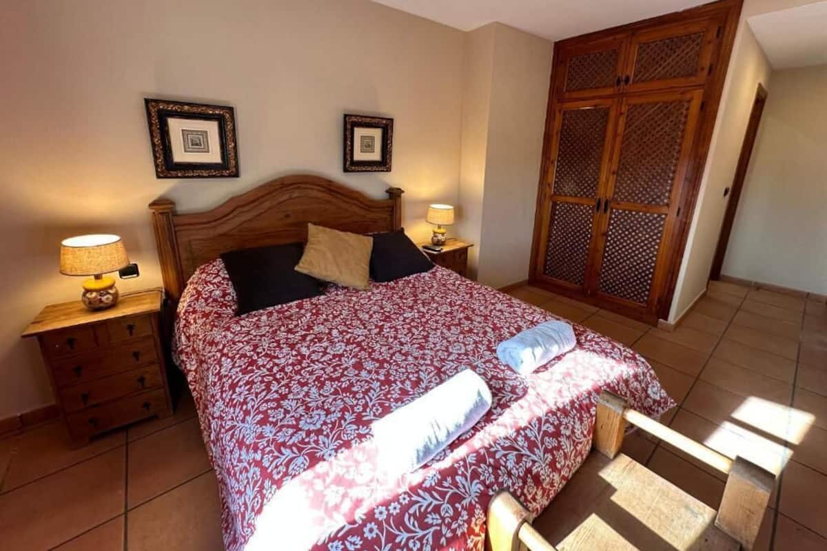 Un dormitorio con cama, cabecero de madera, dos mesitas de noche con lámparas, un armario y un ventilador de techo.