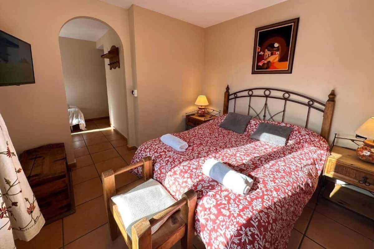 Un dormitorio con una cama doble, una silla de madera, una mesita de noche con lámpara y un cuadro en la pared.