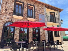 Edificio de piedra con un cartel que indica Hotel Rural y Restaurante Posada La Fragua. Hay mesas y sillas al aire libre con sombrillas rojas.