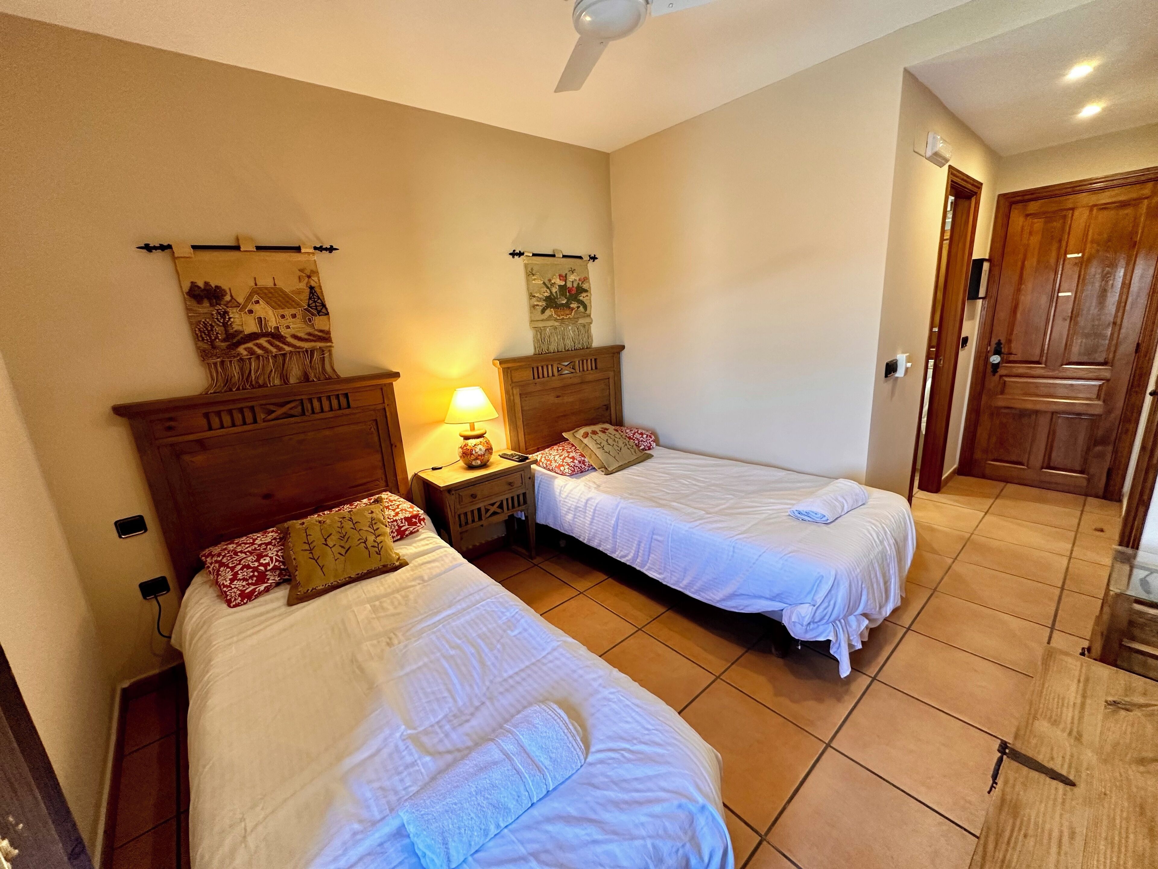 A room with two single beds, wooden headboards, a nightstand with a lamp, and a wall-mounted artwork.