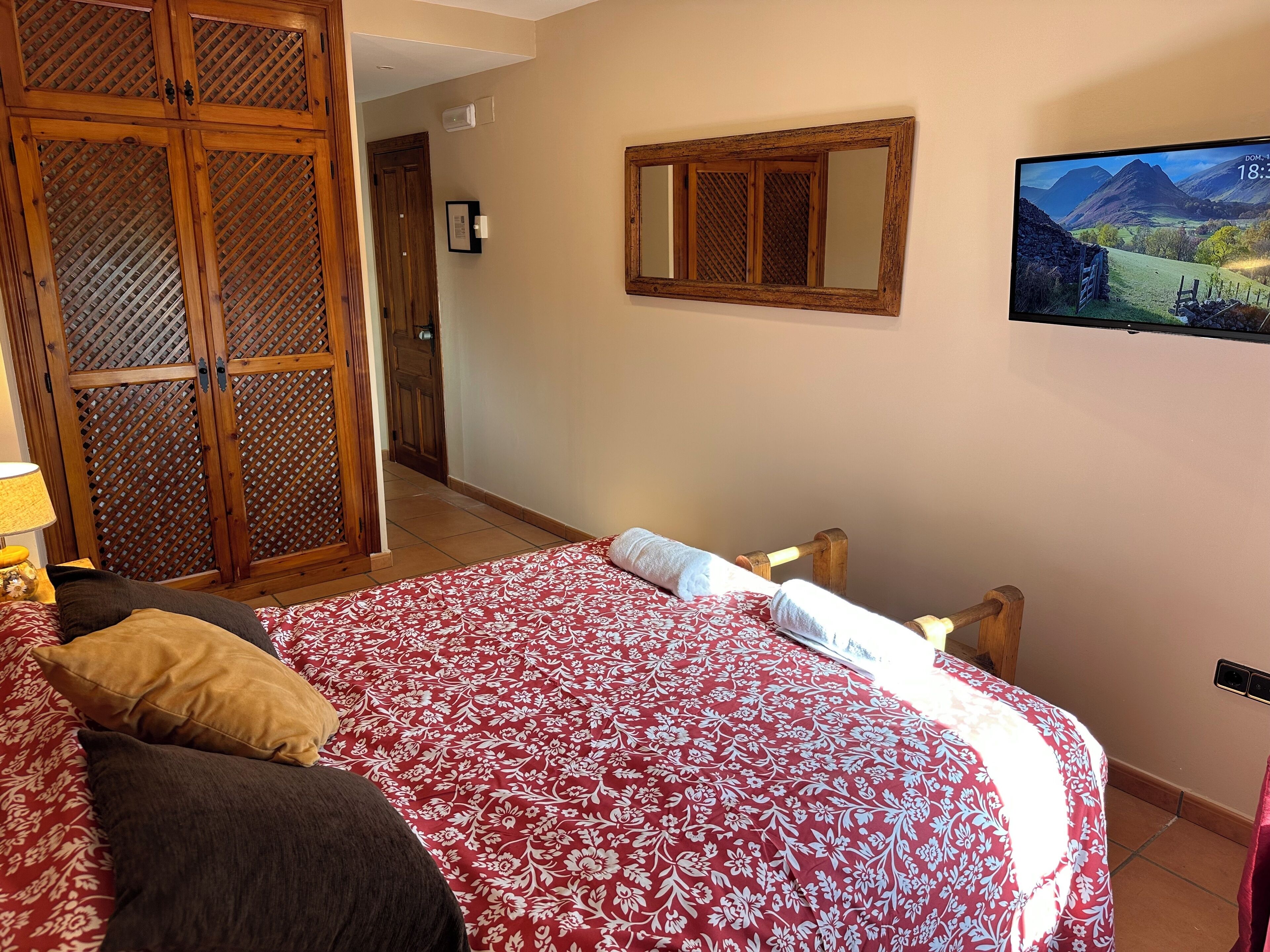 A bedroom with a large bed, a wooden wardrobe, a television mounted on the wall, and a door leading to another room.