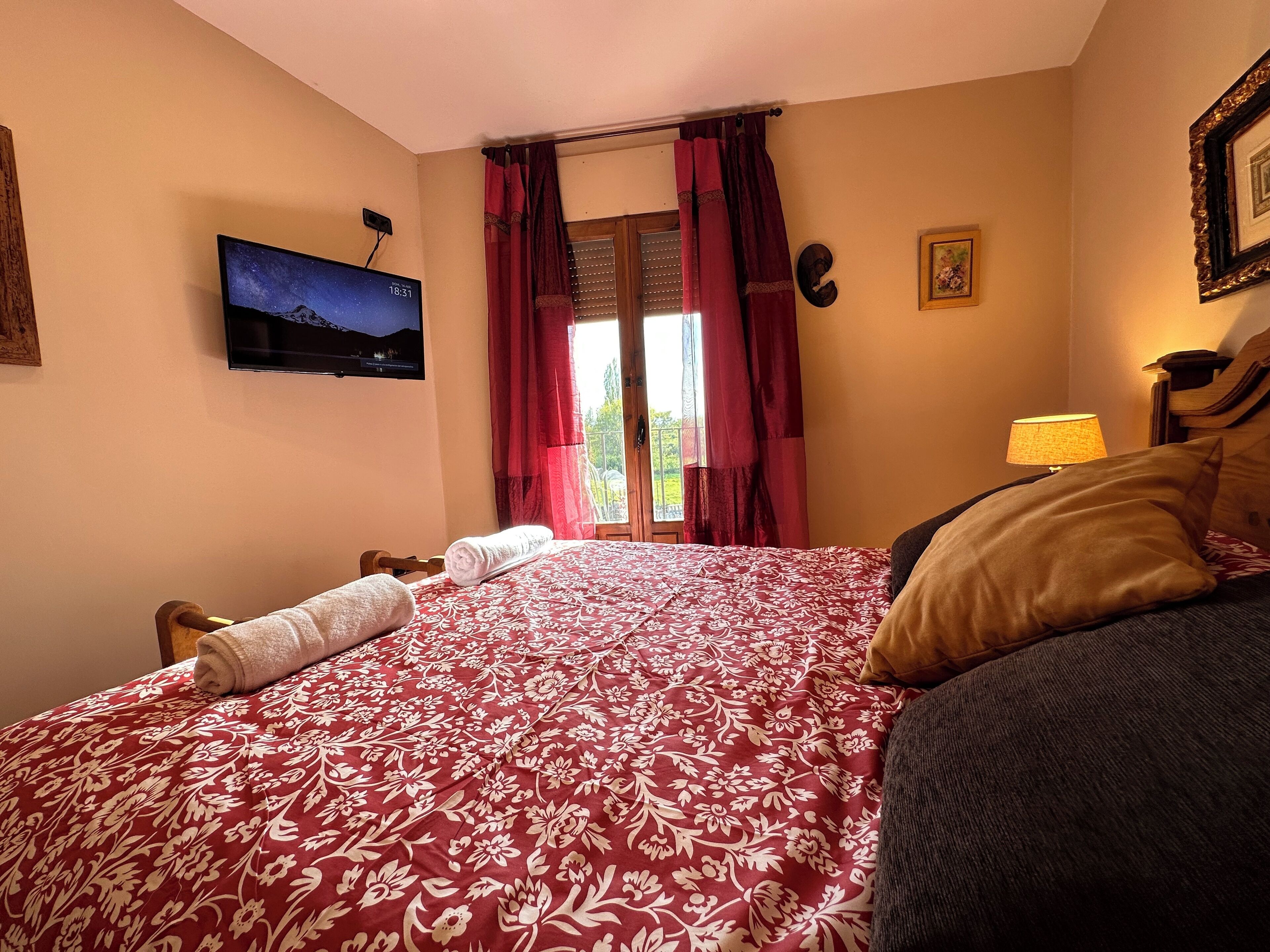 Un dormitorio con cama, televisor montado en la pared, ventana con cortinas y mesita de noche con lámpara.