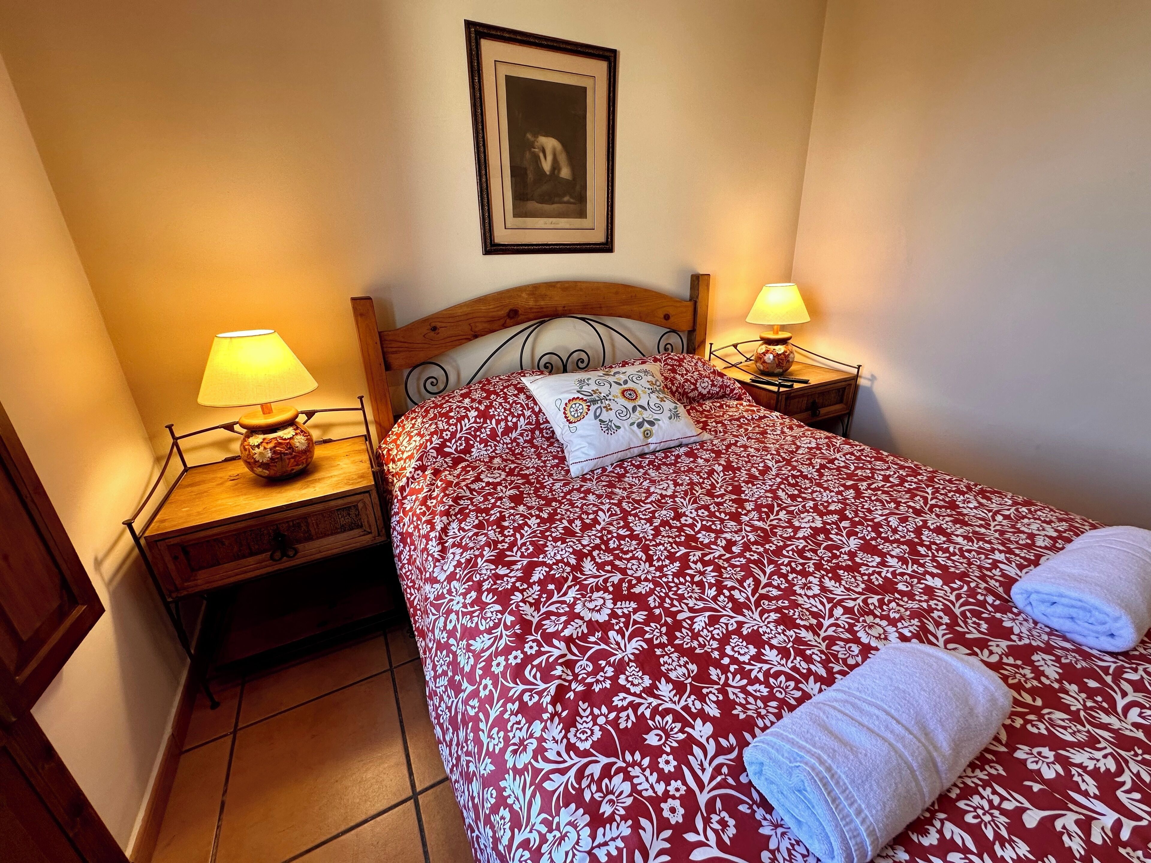 Una cama bien hecha con una colcha estampada con motivos florales, dos lámparas de noche, una mesita de noche de madera y un cuadro en la pared.