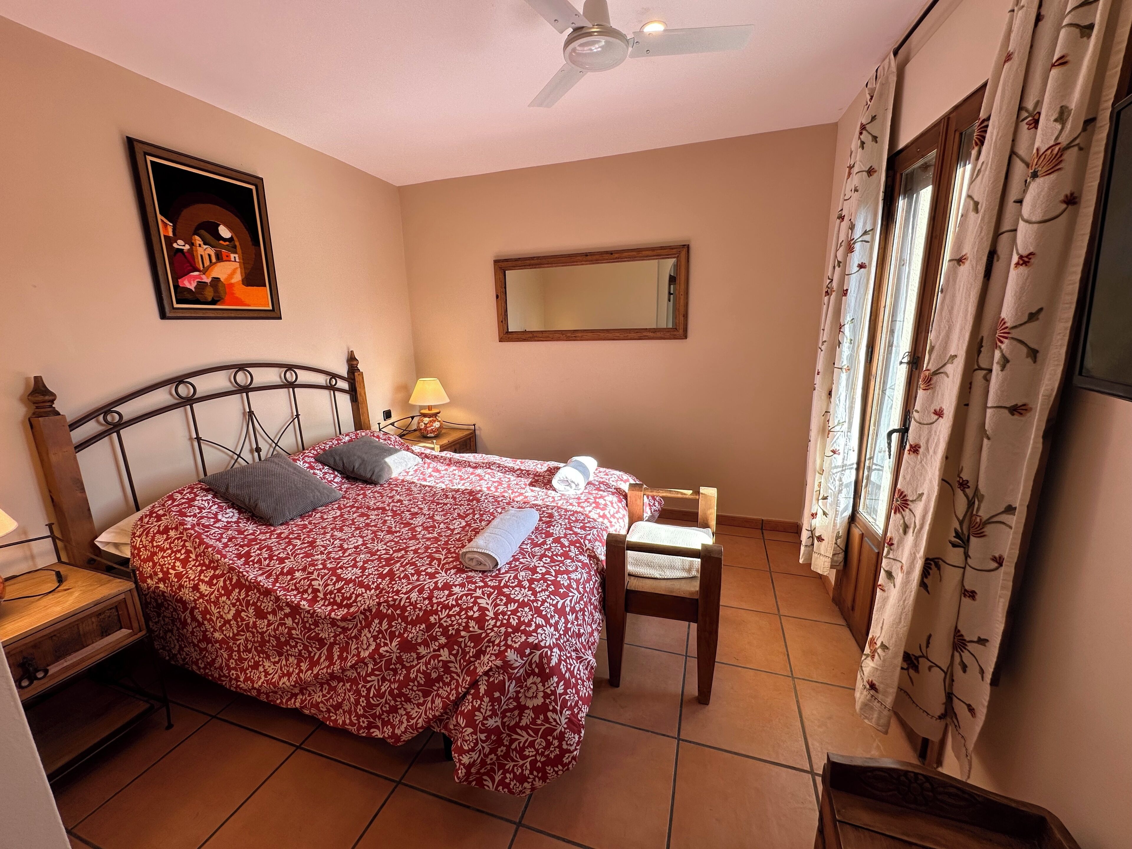 A bedroom with a bed, a chair, a nightstand, a lamp, a ceiling fan, a mirror, and a window with curtains.