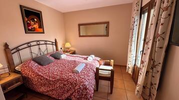 A bedroom with a bed, a chair, a nightstand, a lamp, a ceiling fan, a mirror, and a window with curtains.