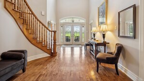 A spacious foyer with a wooden staircase, a grand door with glass panels, a comfortable armchair, and a table with a lamp.