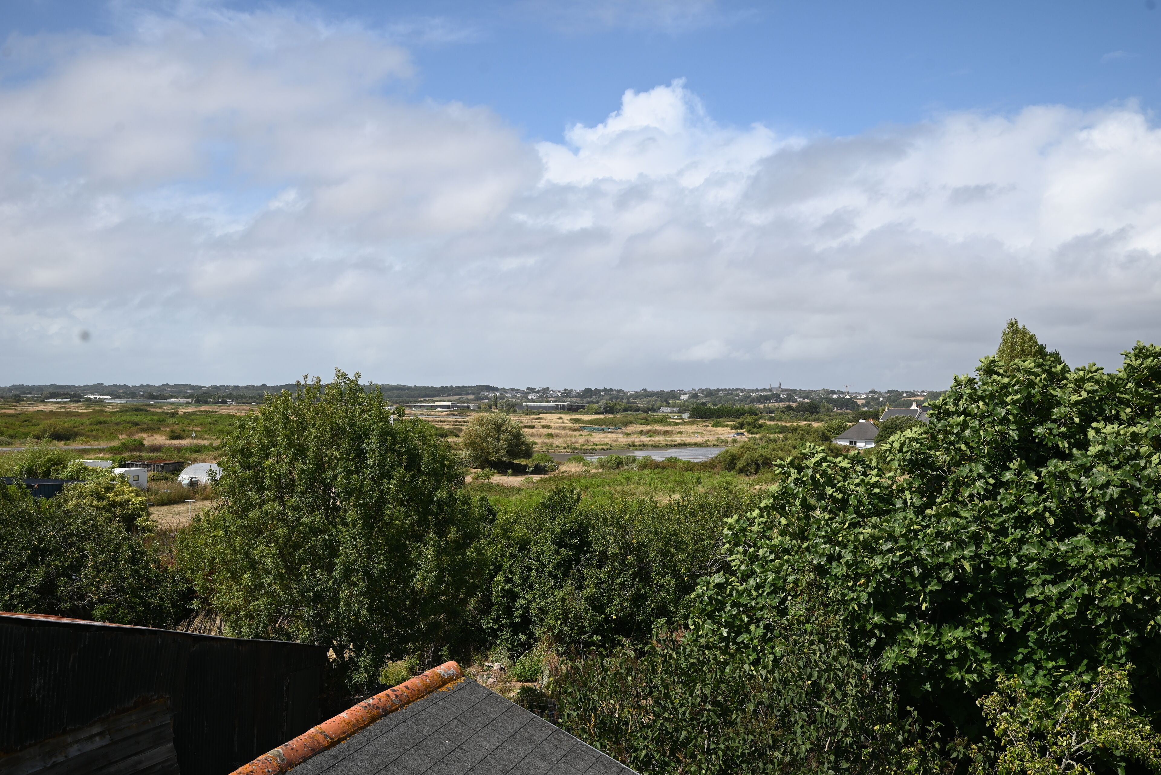 A view from a balcony overlooking a rural landscape with fields, a small body of water, and distant buildings.