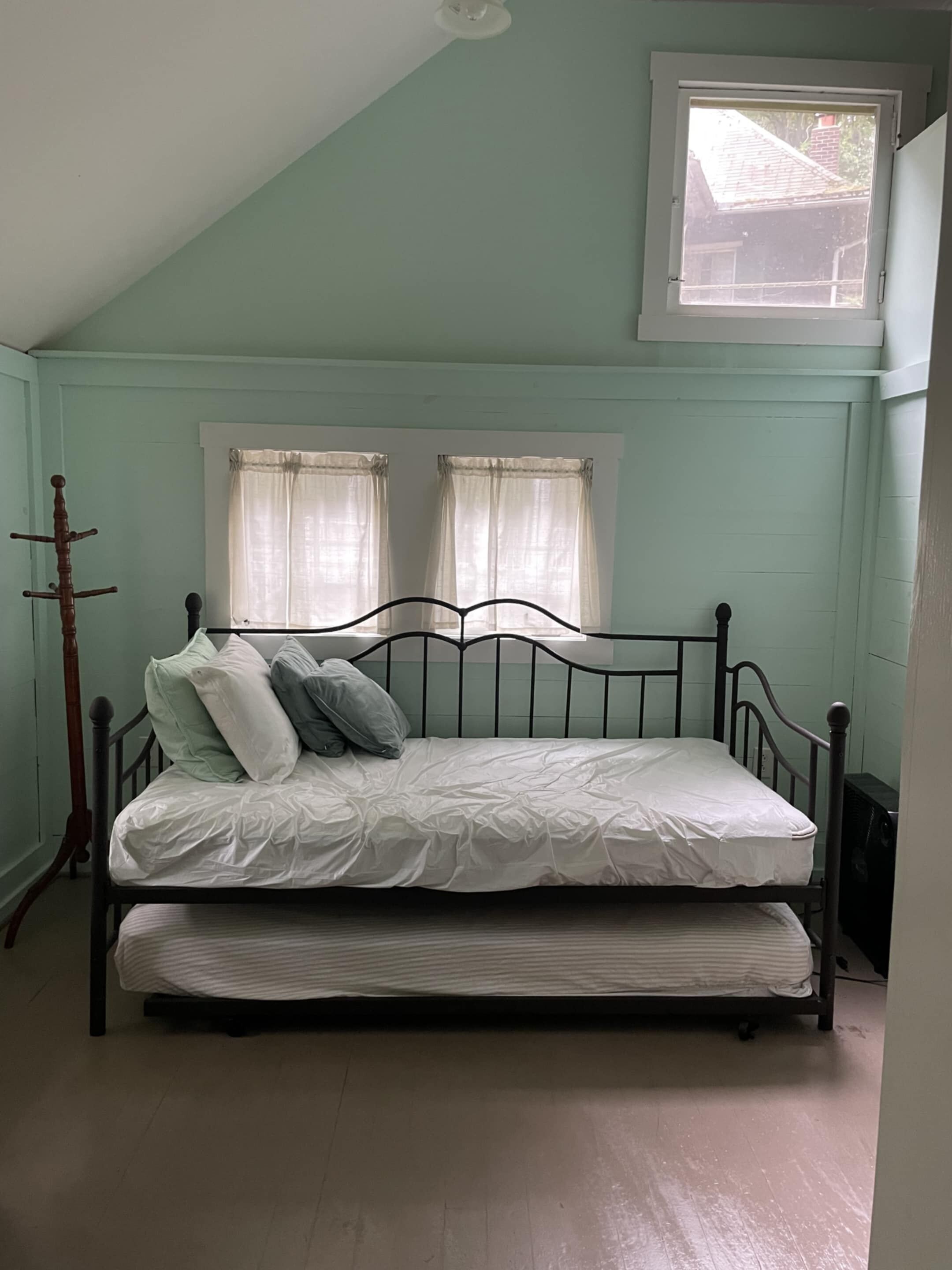 A single bed with a mattress and pillows in a room with light green walls, a sloped ceiling, and two windows with curtains.