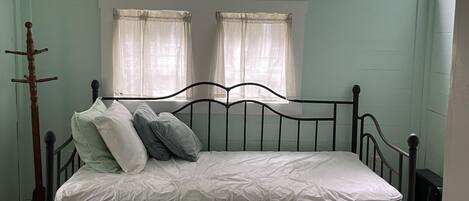 A single bed with a mattress and pillows in a room with light green walls, a sloped ceiling, and two windows with curtains.