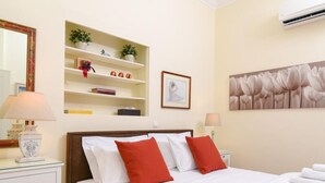 Et hotelværelse med seng, hvide sengetøj, et natbord med lampe, en hylde med pynteting, et indrammet billede og en aircondition.