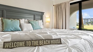 Habitación de hotel con una cama grande, cabecera, vista a la playa y un letrero que dice "WELCOME TO THE BEACH HOUSE".