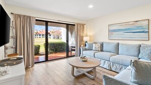 Una sala de estar moderna con un sofá, una mesa de centro y un televisor. Hay un ventanal corredera que da a un balcón con vistas a edificios y zonas verdes.