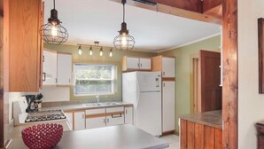 A compact kitchen with white cabinets, a stove, a sink, and a refrigerator. There are two pendant lights hanging above the counter. - Cozy Large House, Big Porch, Fire Pit, Live Music Brewery, Heart of Lake Ann! (Lake Ann)