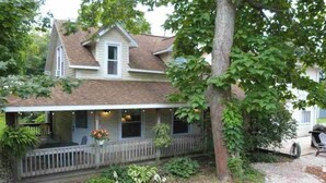 A two-story house with a covered porch and a small garden area. - Cozy Large House, Big Porch, Fire Pit, Live Music Brewery, Heart of Lake Ann! (Lake Ann)