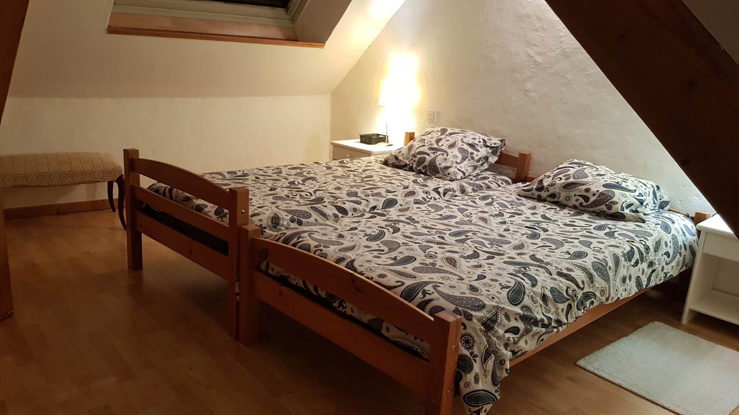 Un dormitorio pequeño y acogedor con cama de madera, ropa de cama estampada, una lámpara de noche y un banco.