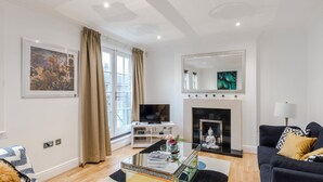 A modern living room with a flat-screen TV, a fireplace, a sofa, and a coffee table with magazines. - Closetoharrods - Closetohydepark - 2 bed House (London)