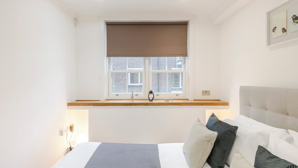 A bedroom with a bed, a window with a view of a brick building, a bedside table, and a wall-mounted shelf. - Closetoharrods - Closetohydepark - 2 bed House (London)