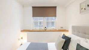 Quarto com cama, janela com vista para um edifício de tijolos, mesa de cabeceira e prateleira na parede.