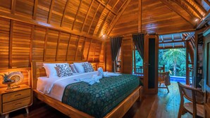 A wooden cabin bedroom with a large bed, a nightstand, and a view of the outdoors. - Villa Mayu (Karangasem)
