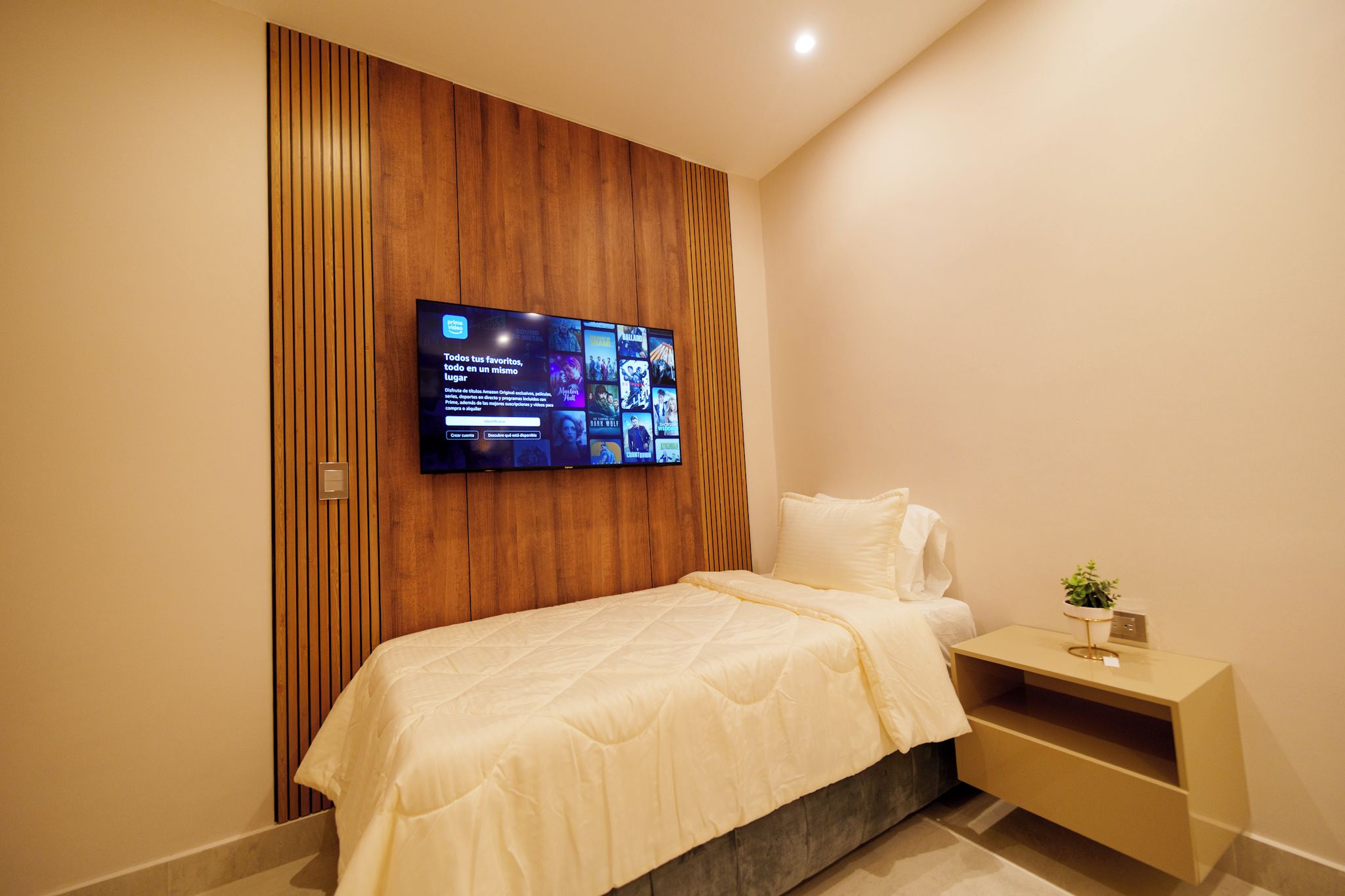 A modern bedroom with a bed, a nightstand, a TV mounted on a wooden panel wall, and a small potted plant.