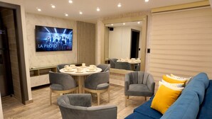 Una camera d'albergo moderna con una TV che mostra 'LA PILOTO', un'area pranzo con tavoli e sedie rotonde e un divano blu con cuscini gialli.
