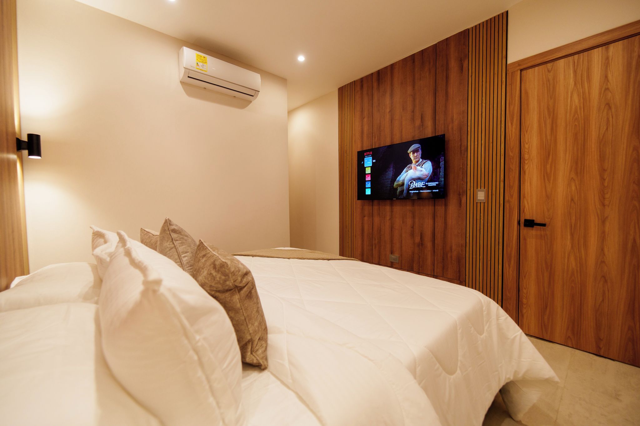 A bedroom with a bed, an air conditioner, a television, and wooden paneling on the wall.