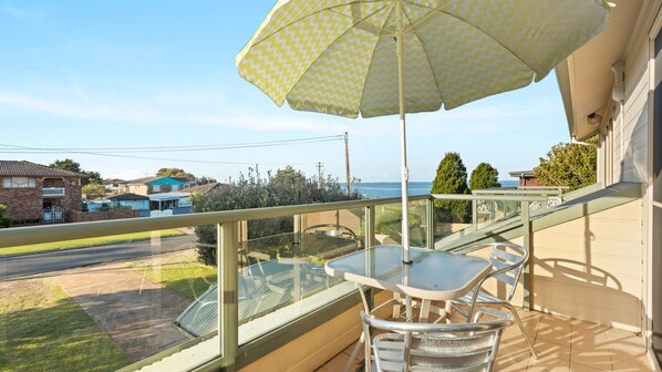Un balcon avec une table et des chaises en verre, un parasol et une vue sur la mer et les maisons voisines.