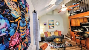A living room with a large floral tapestry, a grey sofa, a wooden dining table, and a kitchen area with wooden cabinets and a black countertop.
