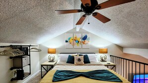 A bedroom with a bed, two bedside tables, a ceiling fan, a wall-mounted lamp, a coat rack, and a painting.