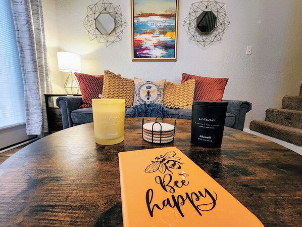 A cozy living room with a wooden coffee table, a yellow candle, a black candle, and a 'Bee Happy' sign.