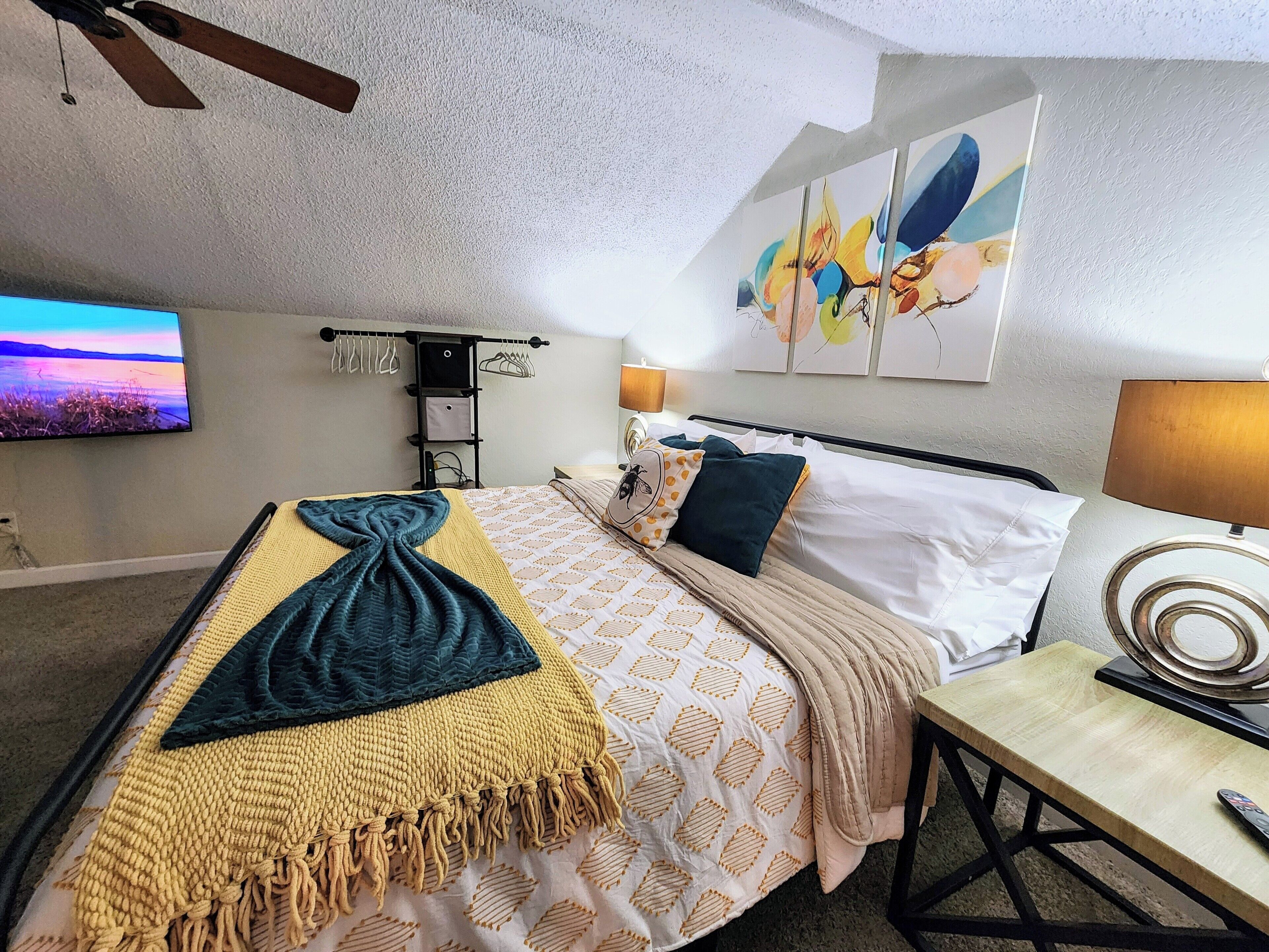 Un dormitorio ordenado con cama, mesita de noche, lámpara y una obra de arte en la pared.