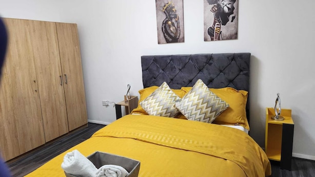 Une chambre avec une grande armoire, un lit avec une literie jaune, deux œuvres d’art accrochées au mur et une table de chevet avec une lampe.