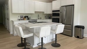 A modern kitchen with white cabinets, a stainless steel refrigerator, a built-in oven, and a marble island with bar stools. - Newly Renovated Lakefront Home with Kayaks and Paddleboards (Locust Grove)