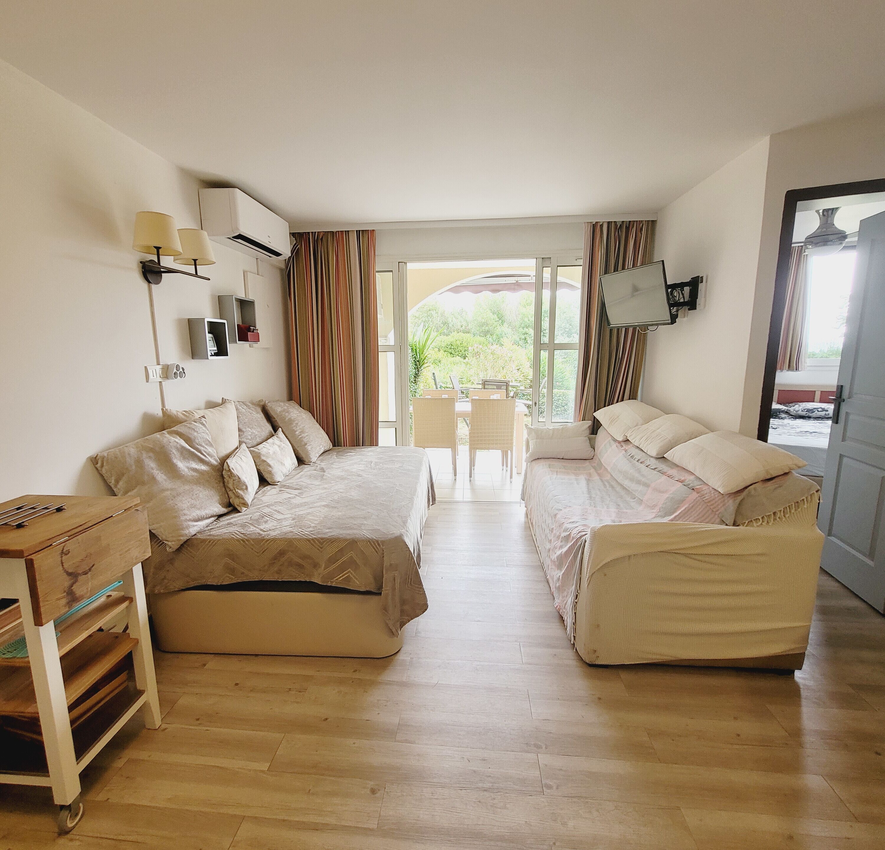 A room with two beds, a desk, and a chair. There is a window with curtains, a TV mounted on the wall, and a door leading outside.