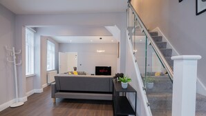 A modern living room with a grey sofa, a black entertainment unit, and a staircase with glass railings. - Stunning 3 Bedroom House-tottenham Stadium-parking (London)