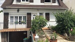 A two-story house with a tiled roof, a front garage, and a small garden area with potted plants and a wooden deck. - Spacious and modern 4 bedroom 2 bathroom House with garden ideal fir families  (Villejuif)