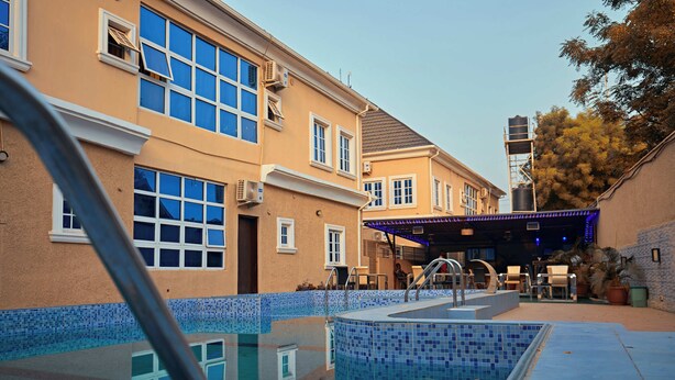 A swimming pool area with a tiled deck, a covered seating area, and a building with multiple windows and air conditioning units. - Apartment 550 (Abuja)