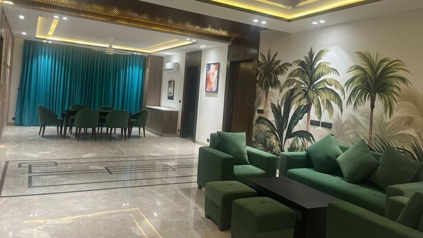 A modern living area with a green sofa set, a dining area with a table and chairs, and a mural of palm trees on the wall. - Tatvam Villa (Sohna)