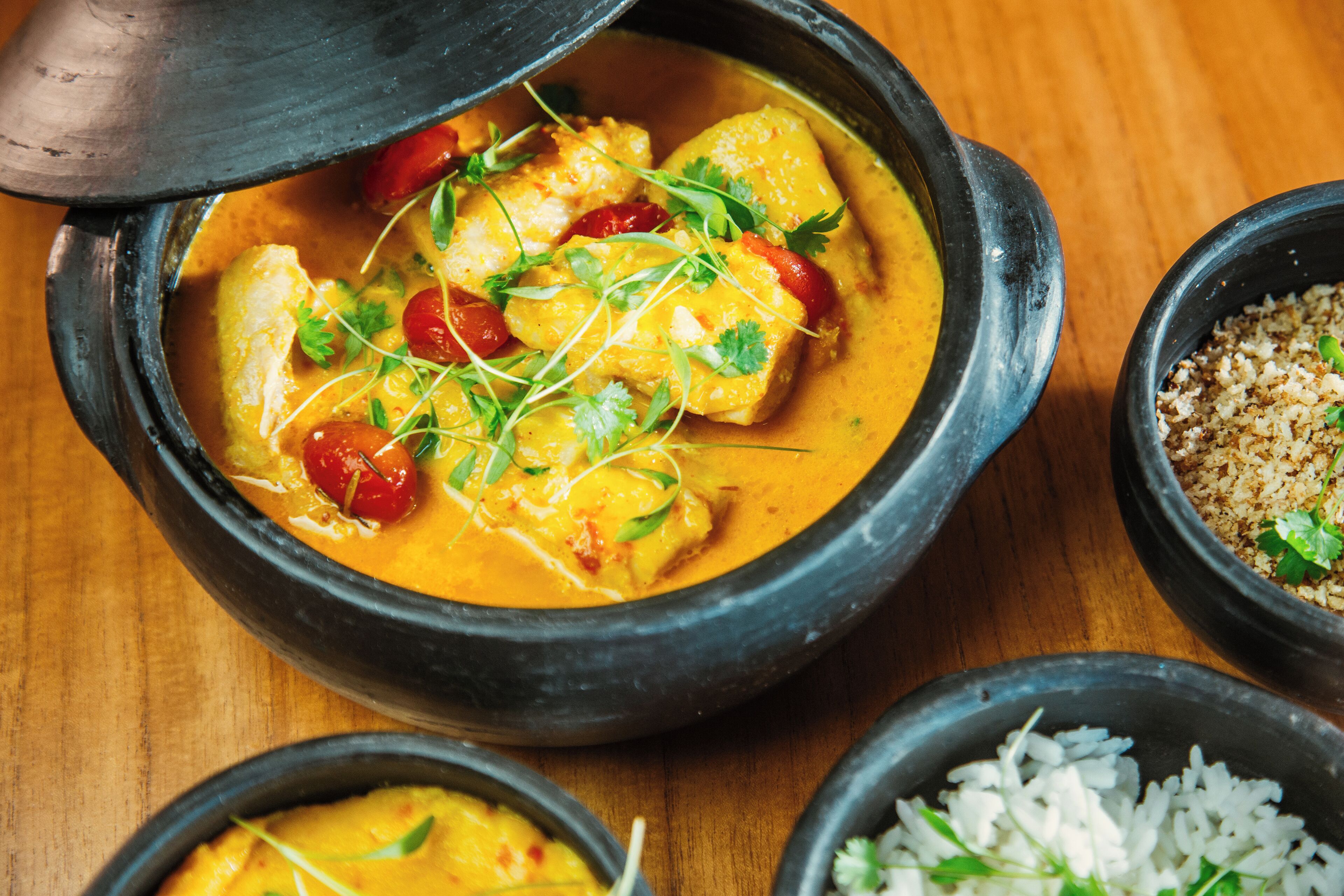 Un pot en terre cuite traditionnel contenant un ragoût, accompagné de bols de riz et d’un petit plat qui semble contenir une sorte de condiment ou un mélange d’épices.