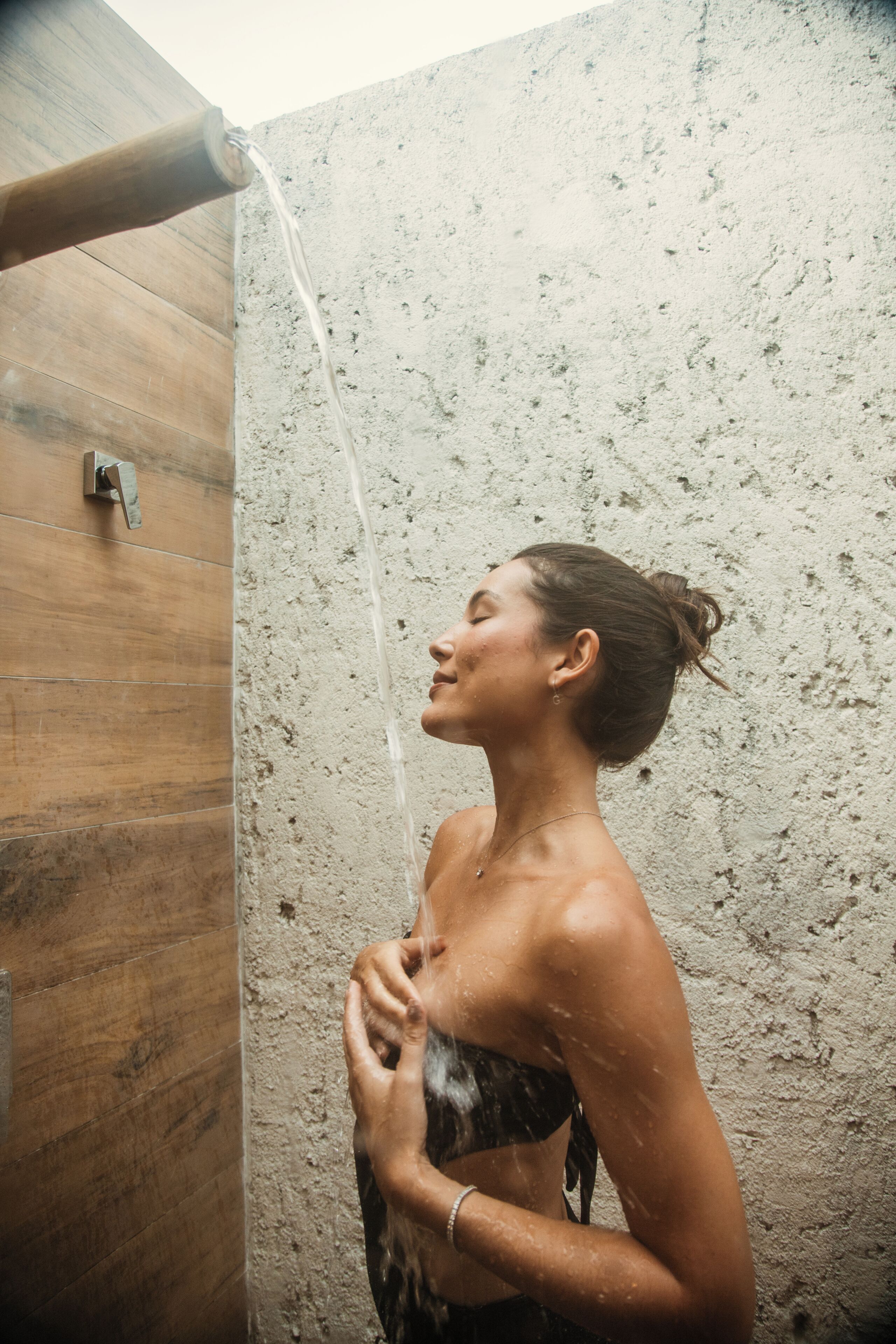 A person in a shower with water flowing, a wooden wall, and a textured wall behind.