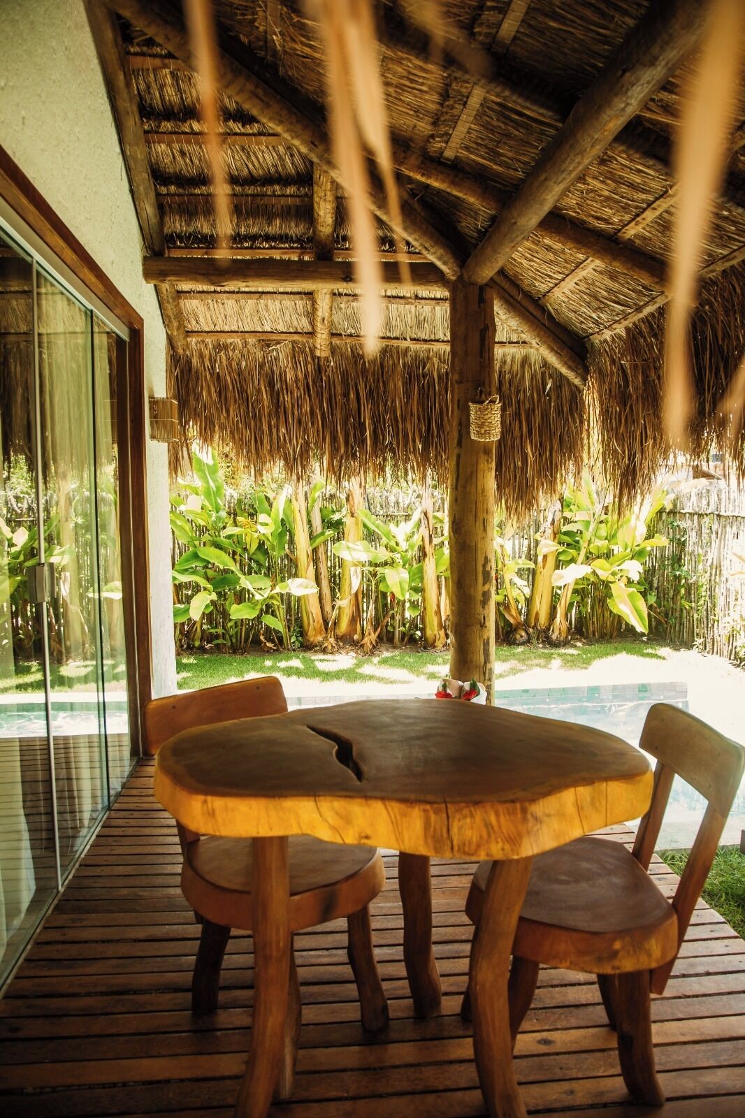 A wooden table with four chairs under a thatched roof, surrounded by lush greenery.
