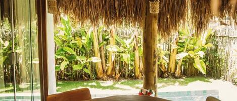 A wooden table with four chairs under a thatched roof, surrounded by lush greenery.