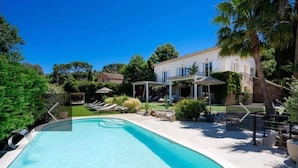Una piscina con sillas de descanso y una pérgola, rodeada de follaje y una casa al fondo.