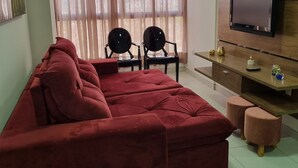 A living room with a red sofa, two black chairs, a wooden TV stand, and a large window with sheer curtains. - Apto Próximo ao Pátio do Forró (Maurício de Nassau Caruaru)