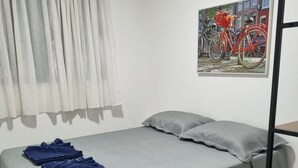 A neatly made bed with a grey bedspread and pillows, a blue towel, and a black rug on a tiled floor. - Apto Próximo ao Pátio do Forró (Maurício de Nassau Caruaru)