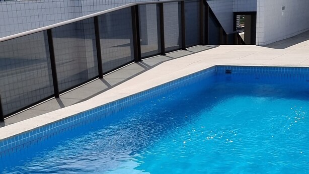 A modern swimming pool with clear blue water, surrounded by a tiled deck and a glass railing. - Apto Próximo ao Pátio do Forró (Maurício de Nassau Caruaru)