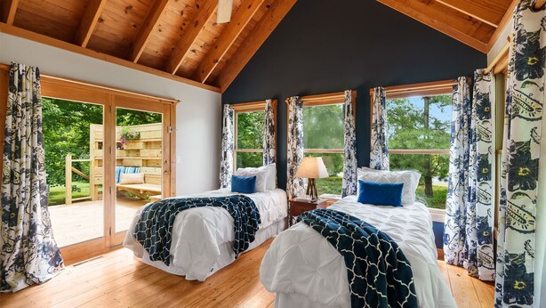 Une chambre avec deux lits, un plancher en bois, de grandes fenêtres avec des rideaux et une vue sur l’extérieur.
