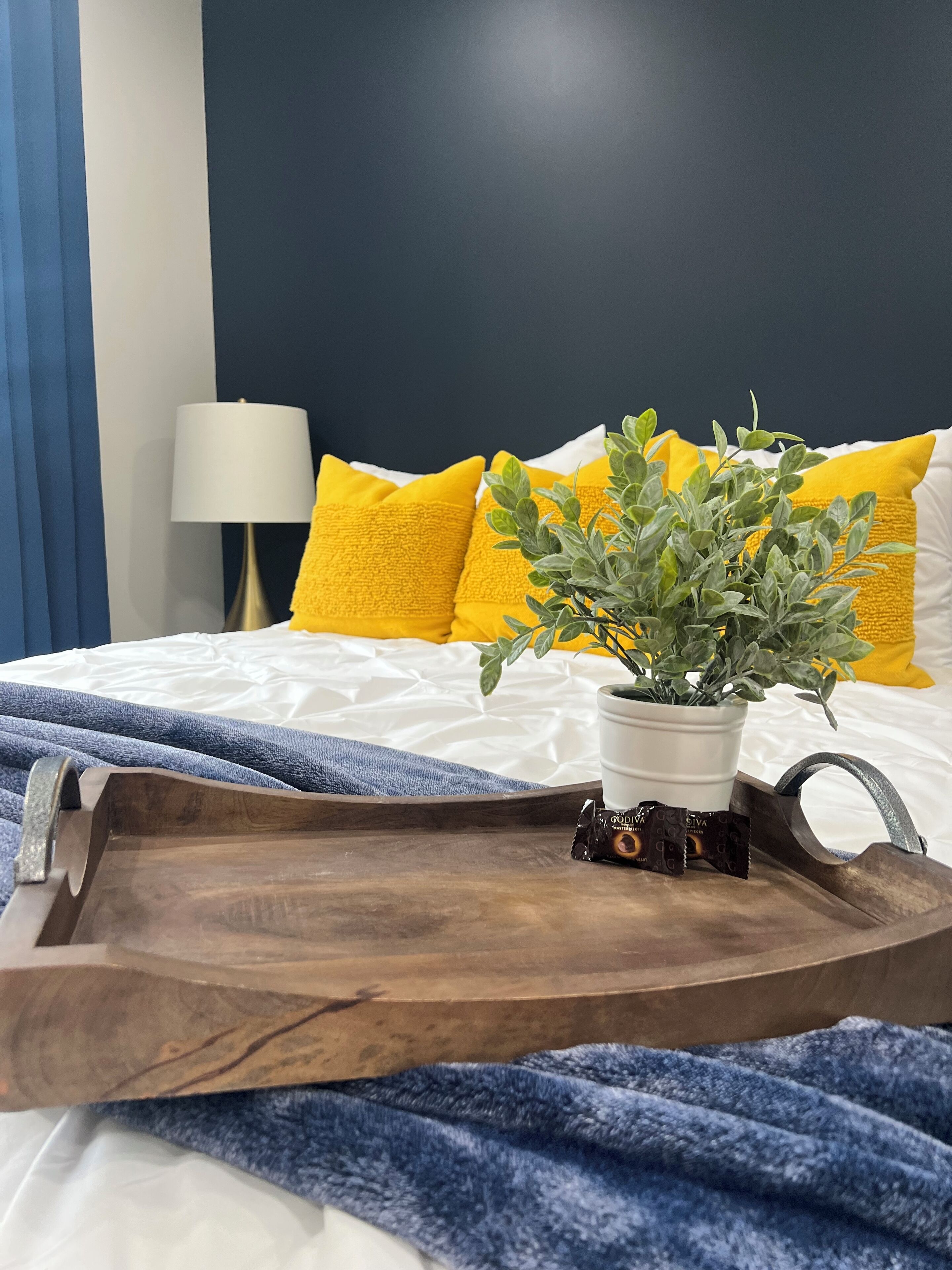 A neatly made bed with a white comforter, a blue and white striped blanket, and a wooden tray with a potted plant and a chocolate bar.