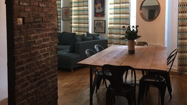 A dining area with a wooden table and chairs, a brick wall, and a green sofa in the background.