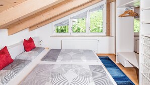 A bedroom with a sloped ceiling, a bed with patterned bedding, a radiator, a window with a view of greenery, and a white wardrobe.